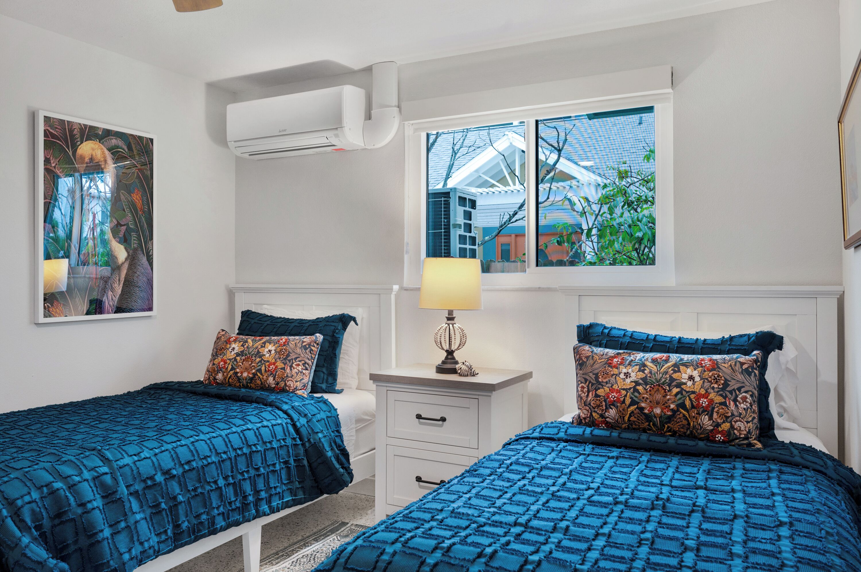 Een slaapkamer met twee bedden, een nachtkastje met een lamp, een raam met uitzicht op gebouwen en een airconditioning.