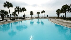Uma piscina grande e cristalina com espreguiçadeiras e palmeiras, cercada por uma grade e com vista para o oceano.