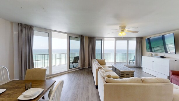 Uma sala de estar espaçosa com um conjunto de sofás, uma mesa de jantar e uma televisão. Grandes janelas proporcionam vista para o oceano.