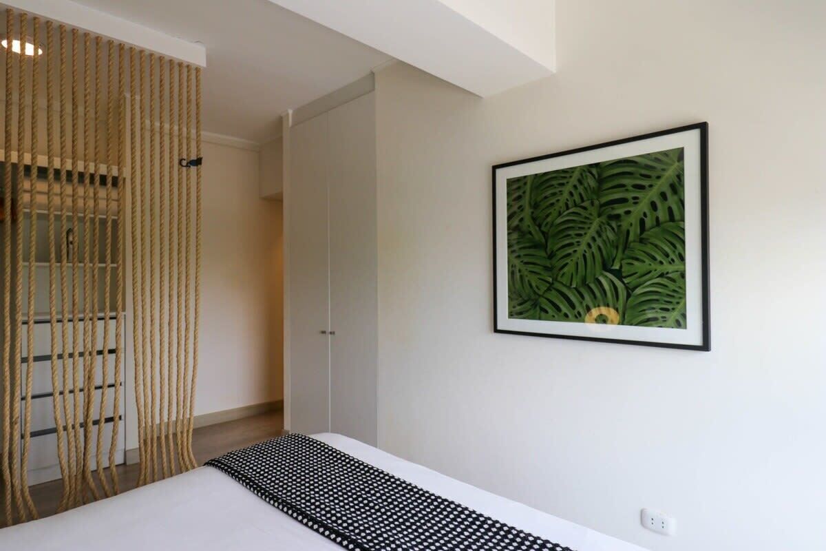 A bedroom with a bed, a framed tropical leaf artwork, and a rope curtain.
