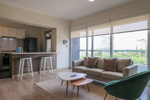 A modern apartment with a kitchen, a living area with a sofa and a green chair, and large windows offering a city view. - w* | Lovely 1BR with Park View in Miraflores (Cercado de Lima)