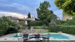 Un espace aménagé au bord de la piscine, avec une table et des chaises, entouré d’une végétation luxuriante et d’un mur en pierre.