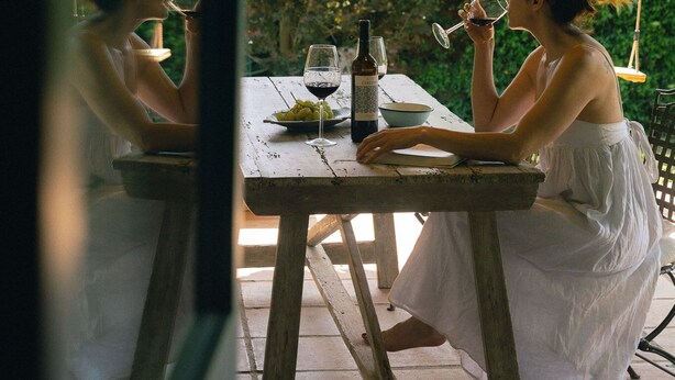 Une femme est assise à une table en bois, devant une bouteille de vin et un verre. Des guirlandes lumineuses vertes sont suspendues au-dessus d’elle, et un mur verdoyant luxuriant se trouve en arrière-plan.