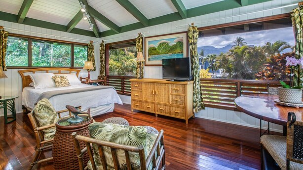Un dormitorio con cama, televisor, armario de madera, silla de mimbre, mesa redonda y ventanas amplias con vistas a palmeras y montañas.