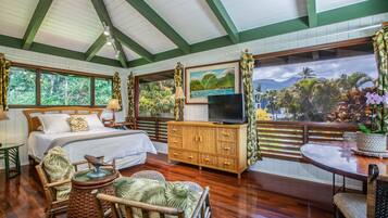 A bedroom with a bed, a TV, a wooden cabinet, a wicker chair, a round table, and large windows with a view of palm trees and mountains.