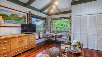 A room with a wooden cabinet, a television, a dining table with chairs, and a view of greenery through a window.