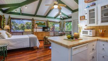 A room with a bed, a dining table, a kitchen area, and a large window with a view of greenery.