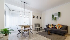 A living room with a sofa, two chairs, a dining table with chairs, a coffee table, a potted plant, and a window with curtains. - MalagaSuite Cathedral Center (Málaga)