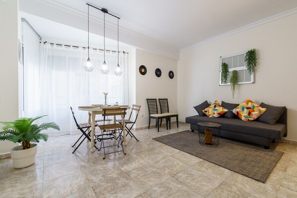 A living room with a sofa, two chairs, a dining table with chairs, a coffee table, a potted plant, and a window with curtains.