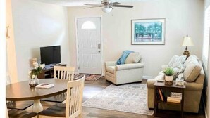 A living room with a dining table, chairs, a sofa, a coffee table, a ceiling fan, and a painting on the wall. - Cottage 304, 2 Bed w/ Fire Pit, near trails! (West Logan)