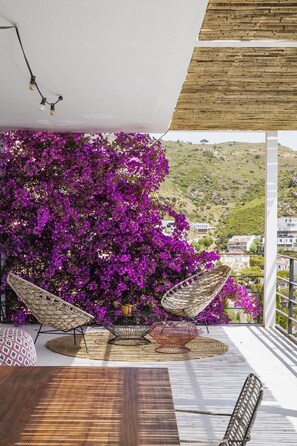 Een balkon met uitzicht op een heuvel, met een rieten stoel, een geweven tafeltje en paarse bougainvillea's.