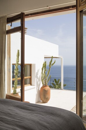 Een slaapkamer met een groot bed, uitzicht op zee vanaf een balkon, en een cactussplant.