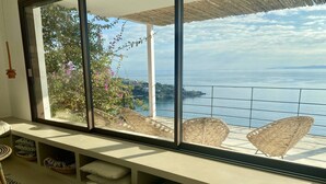 A room with a large window overlooking a coastal view, a built-in bench with cushions, and a wicker chair. - Exclusive & stylish villa. Concierge service included (Roses)