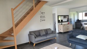 A modern living room with a grey sofa, a blue armchair, a wooden staircase, and a kitchen area in the background.