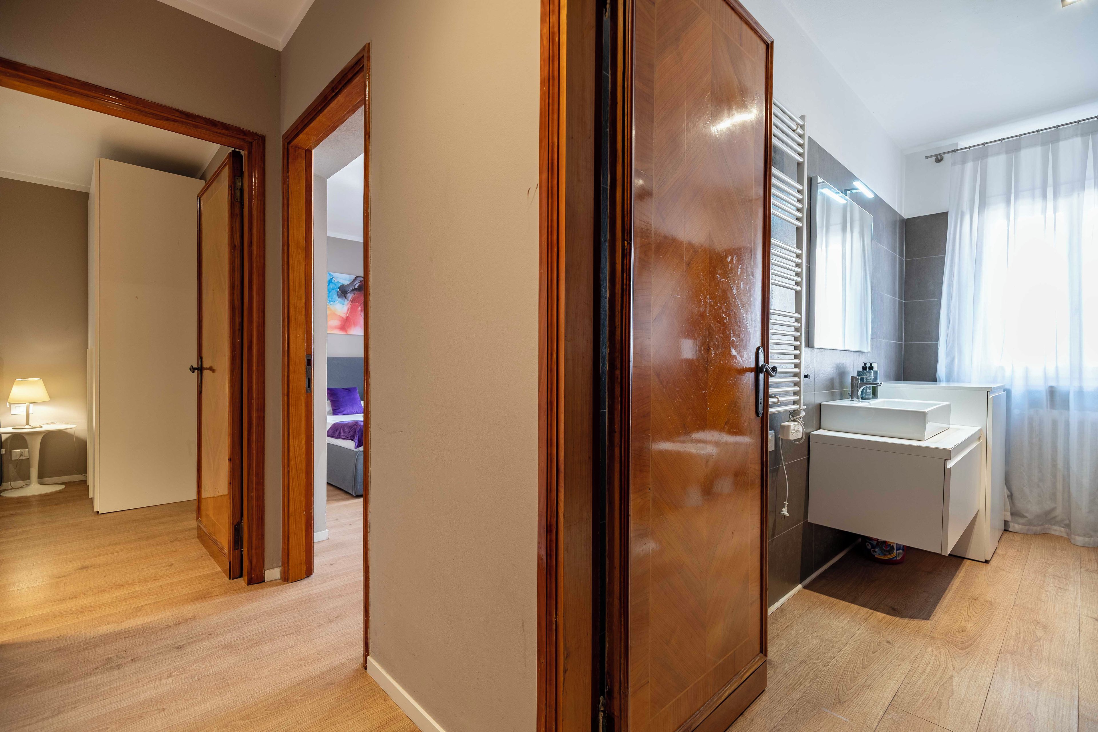 Une chambre d’hôtel avec une armoire en bois, une salle de bain avec un lavabo blanc et un lit avec un oreiller violet.
