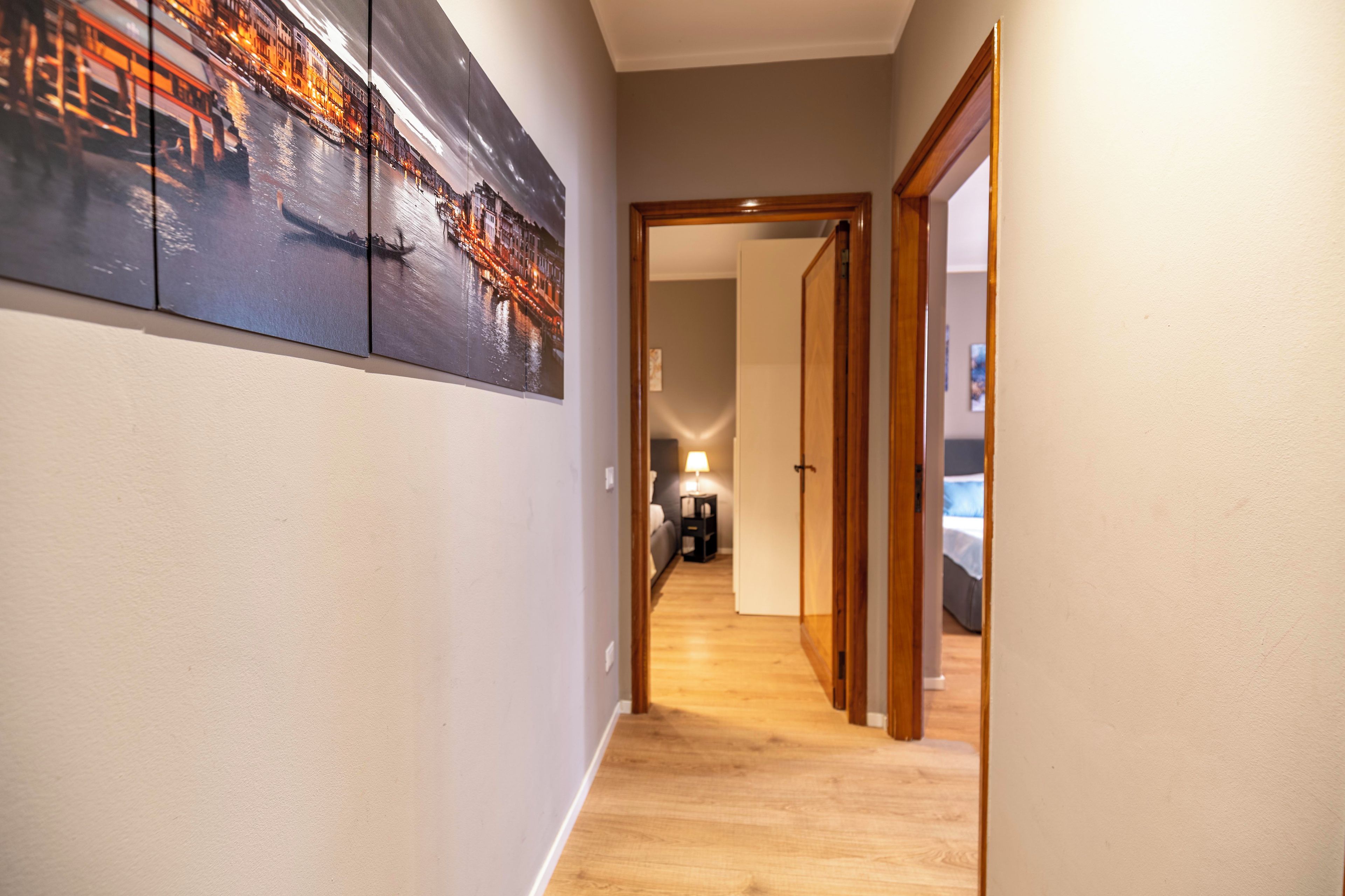 A hallway with a cityscape painting on the wall, wooden flooring, and a door leading to a bedroom.