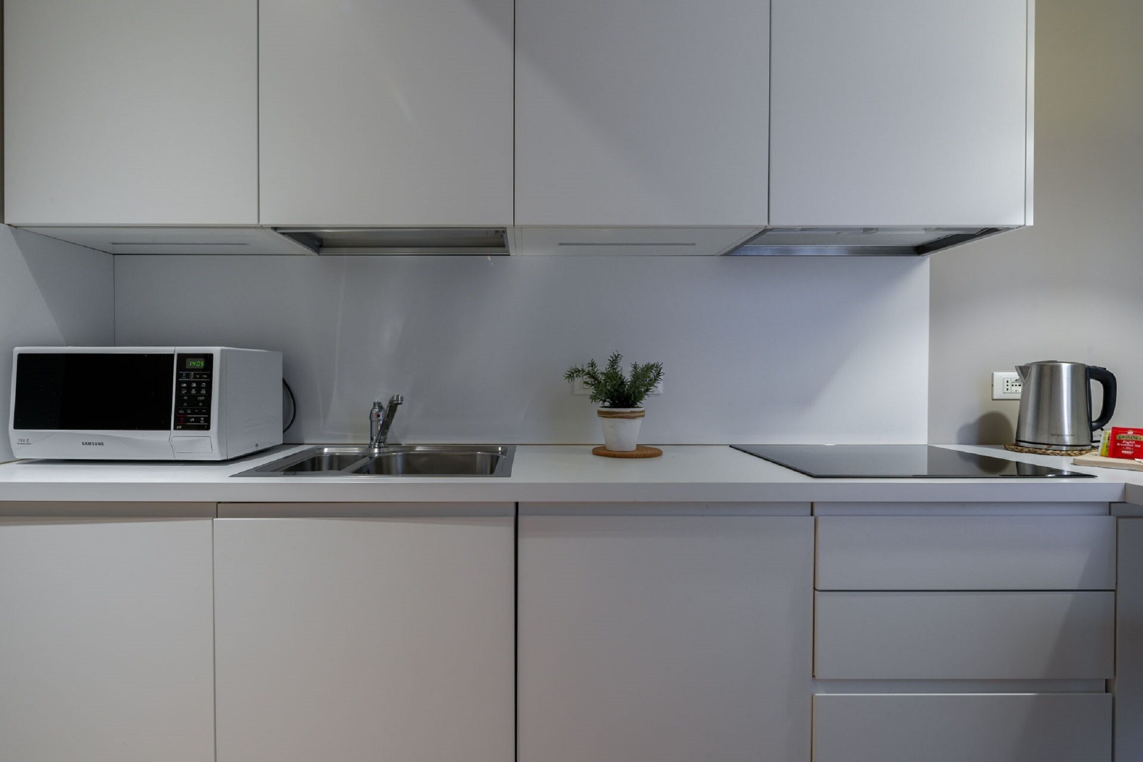 Une cuisine moderne avec des meubles blancs, un four à micro-ondes, un évier, une petite plante en pot et une bouilloire.