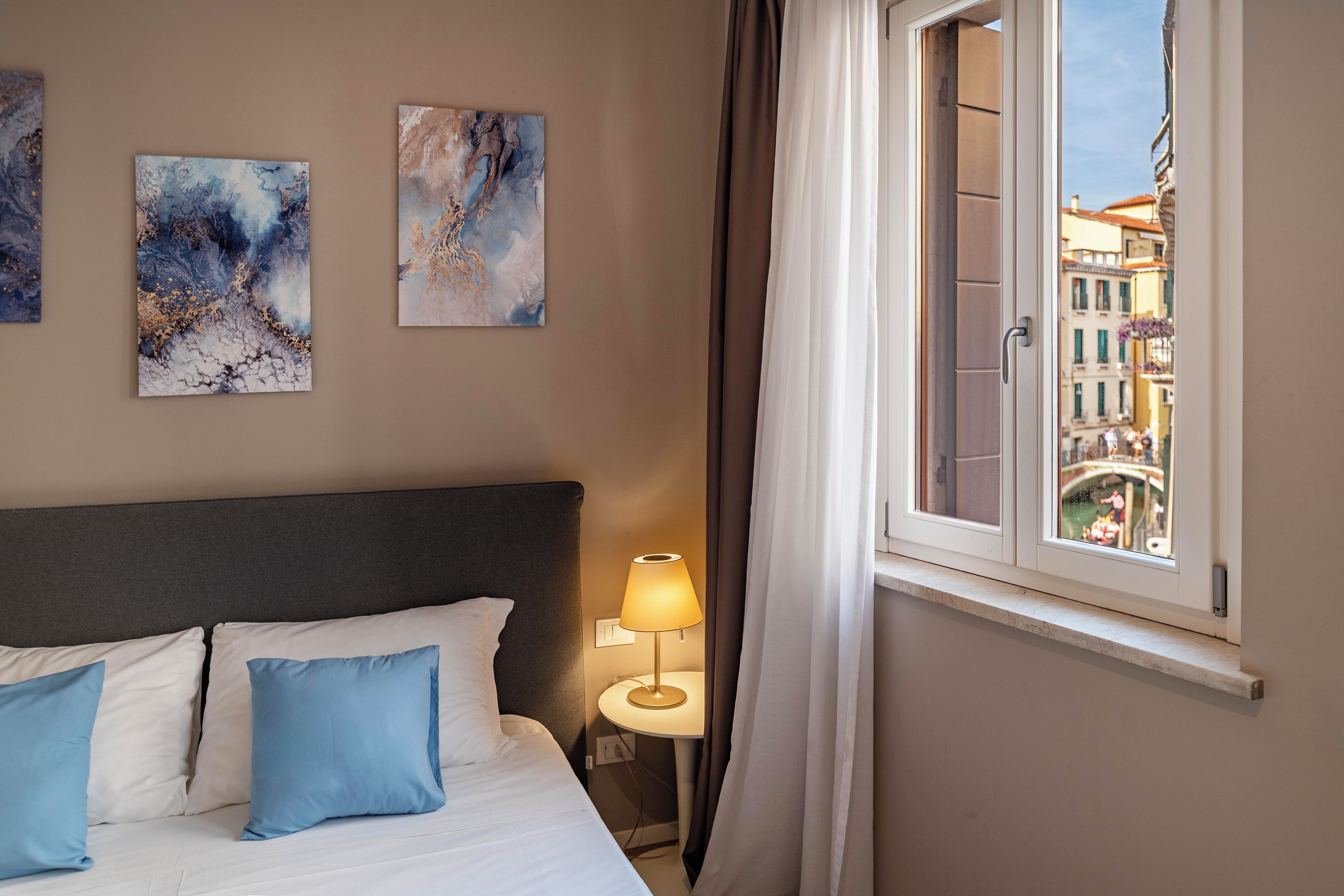 A hotel room with a bed, pillows, a lamp, and abstract art on the wall.
