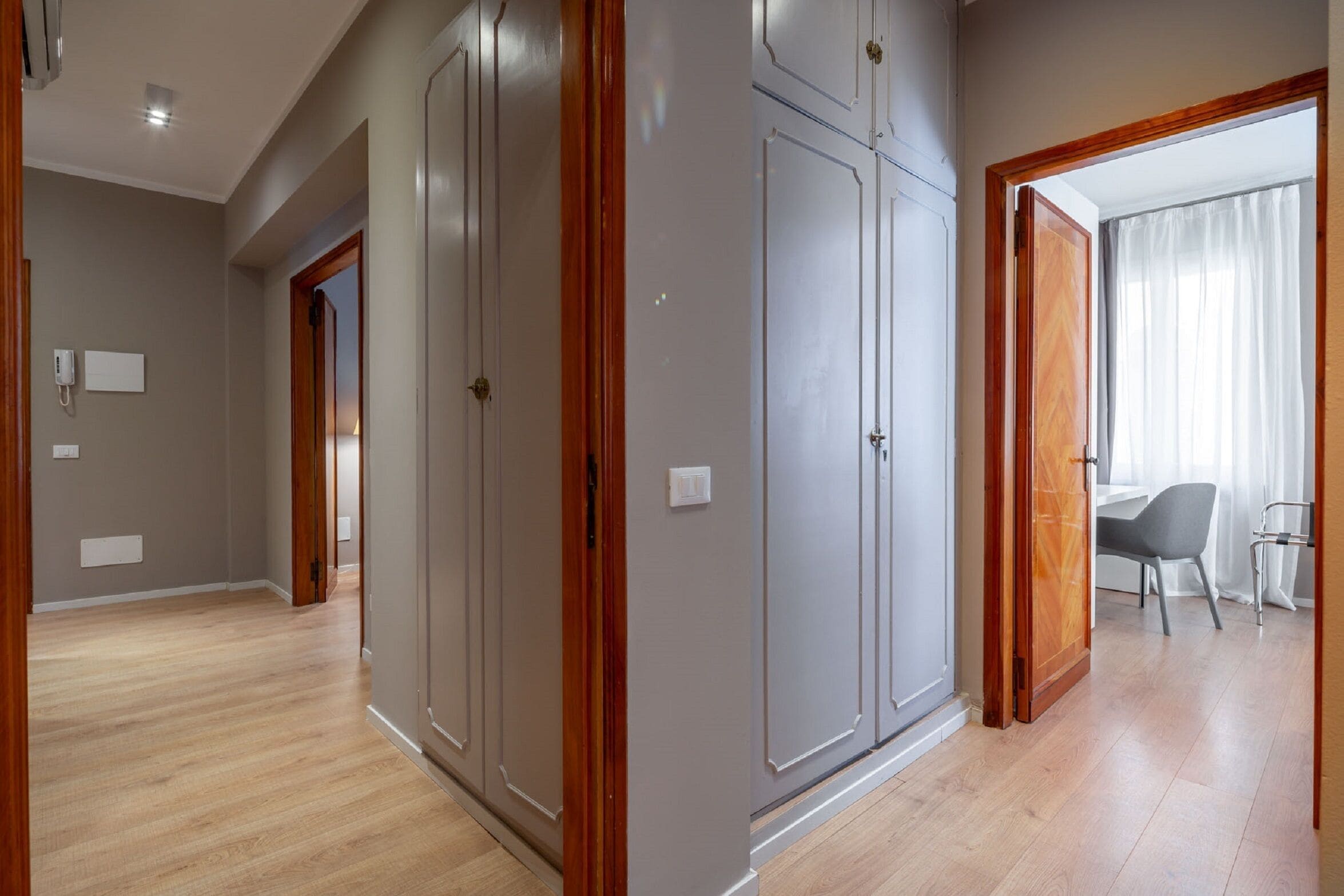 Un couloir doté d’un plancher en bois, de murs gris et d’une porte donnant sur une pièce où se trouvent un bureau et une chaise.