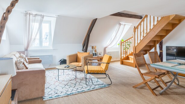 En rummelig loftrum med højt til loftet, en trætrappe, en sofa, en lænestol og et spisebord.