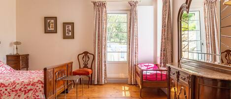 A bedroom with a wooden floor, a bed with a floral bedspread, a chair, a dresser, and a large mirror.