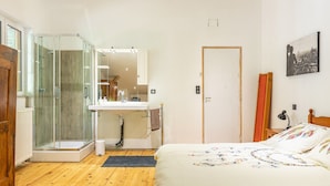 Un dormitorio con piso de madera, una cama con colcha colorida, una mesita de noche blanca, un espejo, una ducha con puerta de vidrio y una puerta con cortina roja.