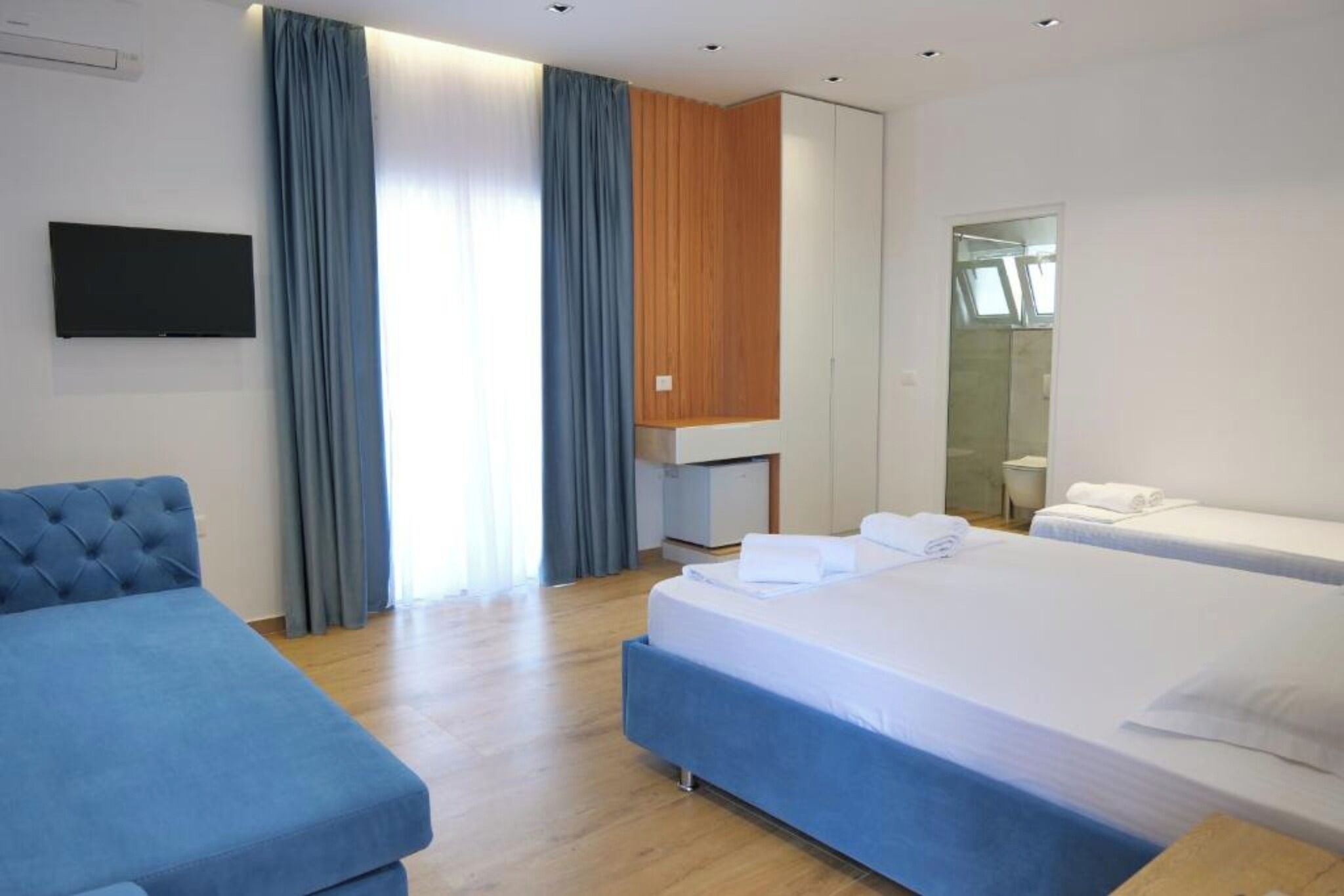 Camera d'albergo con un letto grande, un letto più piccolo, un divano, una televisione, una parete rivestita in legno e un bagno visibile attraverso una porta in vetro.