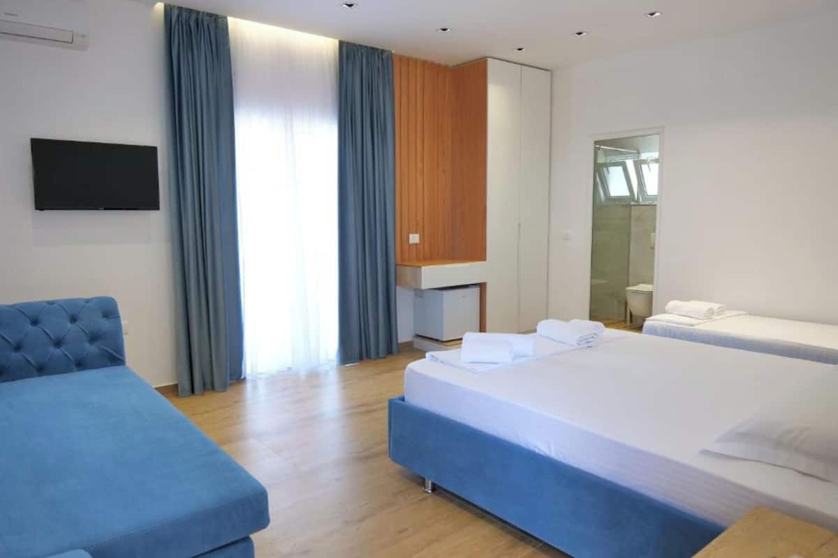 Ein Hotelzimmer mit einem großen Bett, einem kleineren Bett, einer Schlafcouch, einem Fernseher, einer Wand mit Holzpaneelen und einem durch eine Glastür einsehbaren Badezimmer.