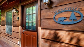 A wooden cabin with a 'WELCOME' sign featuring a bear and a tree, a brown door, and two wall-mounted lanterns. - Cabin in the Woods w/ Hot Tub (Sevierville)