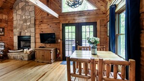 A log cabin interior with a stone fireplace, wooden dining table, and a television. - Cabin in the Woods w/ Hot Tub (Sevierville)