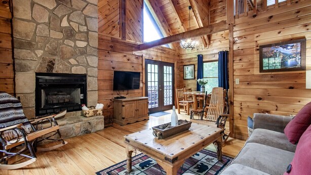 A cozy log cabin interior with a stone fireplace, wooden furniture, and a large window. - Cabin in the Woods w/ Hot Tub (Sevierville)