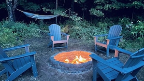 A fire pit with four Adirondack chairs around it, surrounded by greenery. - Cozy Cottage with Pool+Jacuzzi+Sauna+Fire Pit and Reading Nook! (Livingston Manor)