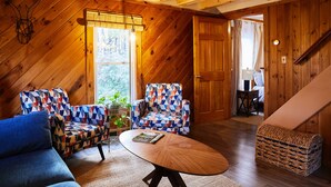 A cozy room with wooden walls and a sloped ceiling, featuring two patterned armchairs, a wooden coffee table, and a wicker basket. - Cozy Cottage with Pool+Jacuzzi+Sauna+Fire Pit and Reading Nook! (Livingston Manor)