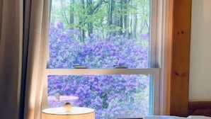 A room with a bed, a nightstand with a lamp, and a window with a view of trees and purple flowers. - Cozy Cottage with Pool+Jacuzzi+Sauna+Fire Pit and Reading Nook! (Livingston Manor)
