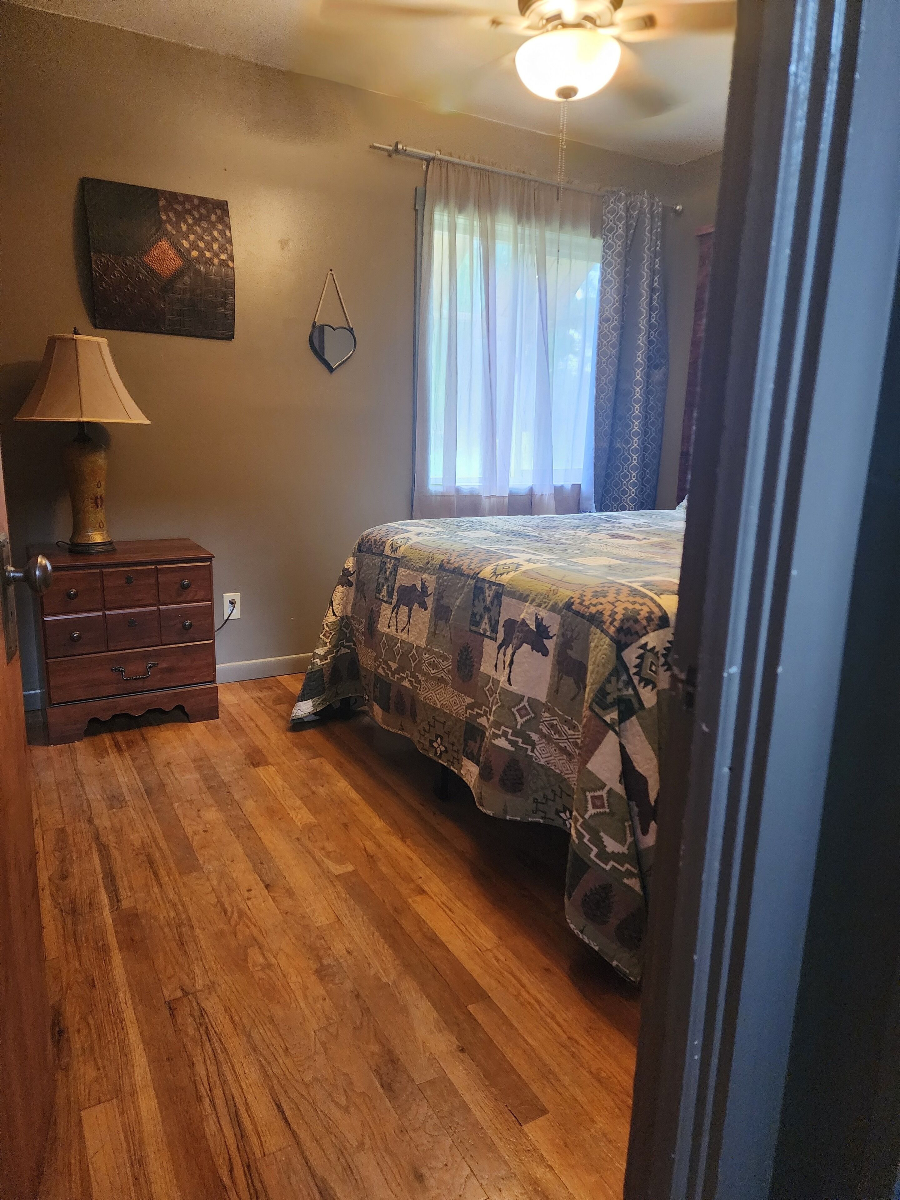 A bedroom with a wooden floor, a bed with a patterned bedspread, a lamp, a dresser, and a ceiling fan.