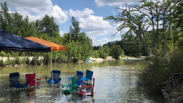 A clear river with chairs partially submerged, a blue canopy, and trees in the background.