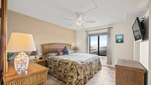 Ein Schlafzimmer mit Bett, Nachttisch mit Lampe, Deckenventilator und an der Wand montiertem Fernseher.