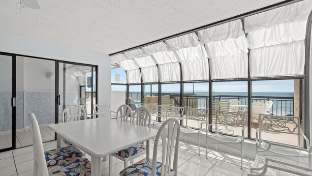 Un comedor con una mesa y sillas blancas, ventanas amplias con cortinas blancas y vista al mar.