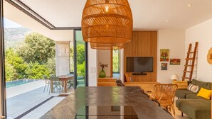 Un salon moderne avec une grande table en verre, un luminaire suspendu en osier, un téléviseur à écran plat et un canapé vert agrémenté de coussins jaunes.
