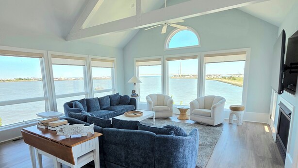 A spacious living room with a large sectional sofa, a coffee table, and two armchairs. There are large windows offering a view of the water and a bridge. - Sunrise-Sunset Views From Waterfront Home w/ Elevator (just renovated) (Bethany Beach)