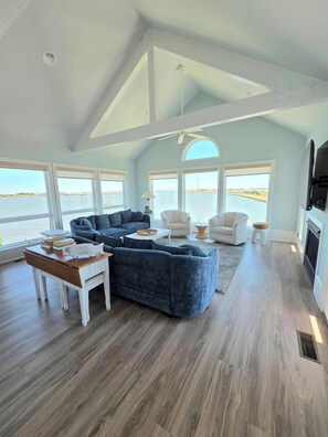 A spacious living room with a large sectional sofa, a coffee table, and two armchairs. There are large windows offering a view of the water and a bridge.