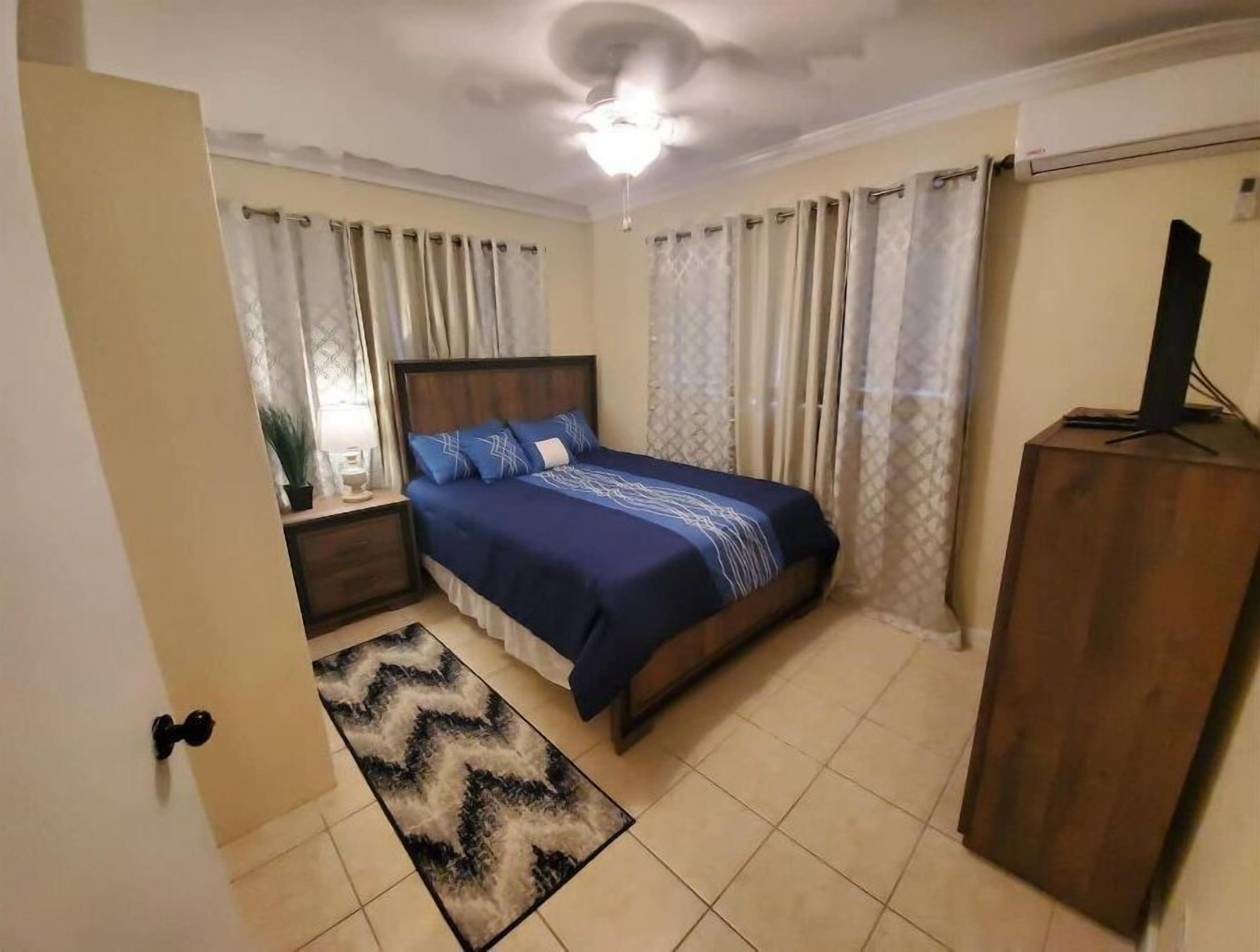 A bedroom with a bed, curtains, a ceiling fan, a TV on a wooden cabinet, and a patterned rug.