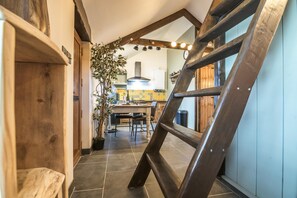 Un pasillo estrecho con una escalera de madera, una planta en maceta y una zona de cocina con salpicadero de azulejos y armarios de madera.