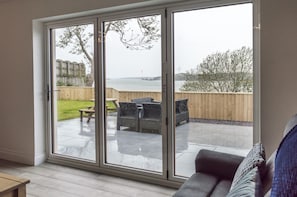 A room with a sliding glass door leading to an outdoor patio with a view of the sea and a wooden fence.