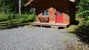 Een klein houten vakantiehuisje met een rode deur, een raam en een veranda met een tafel en stoelen.