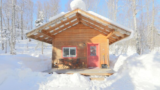 A wooden cabin with a red door, a window, and a porch with chairs.