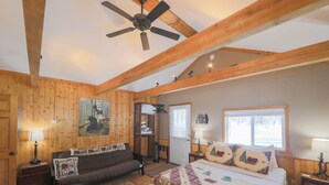 A bedroom with a bed, a sofa, a ceiling fan, wooden beams, a window, and a door.