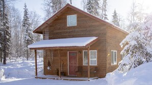 A wooden cabin with a porch, a red door, and a snow-covered landscape. - Willow Cabin (Talkeetna)
