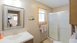 A bathroom with a white sink, a mirror, a shower, and a sign on the wall. - Willow Cabin (Talkeetna)