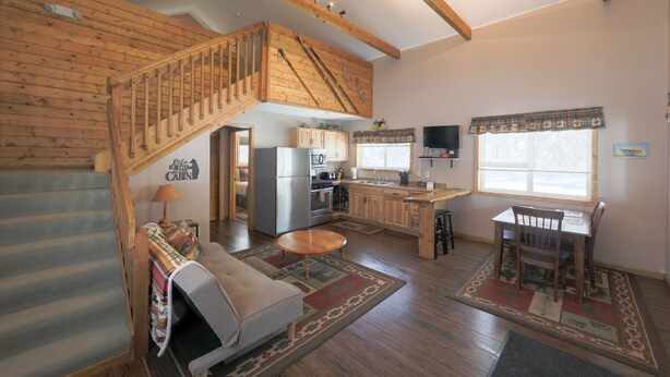 A spacious living area with a wooden staircase, a sofa, a dining table, and a kitchen in the background. - Willow Cabin (Talkeetna)